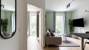Una sala de estar moderna con sofá, mesa de comedor y televisor. Hay ventanas amplias y una planta en la habitación.