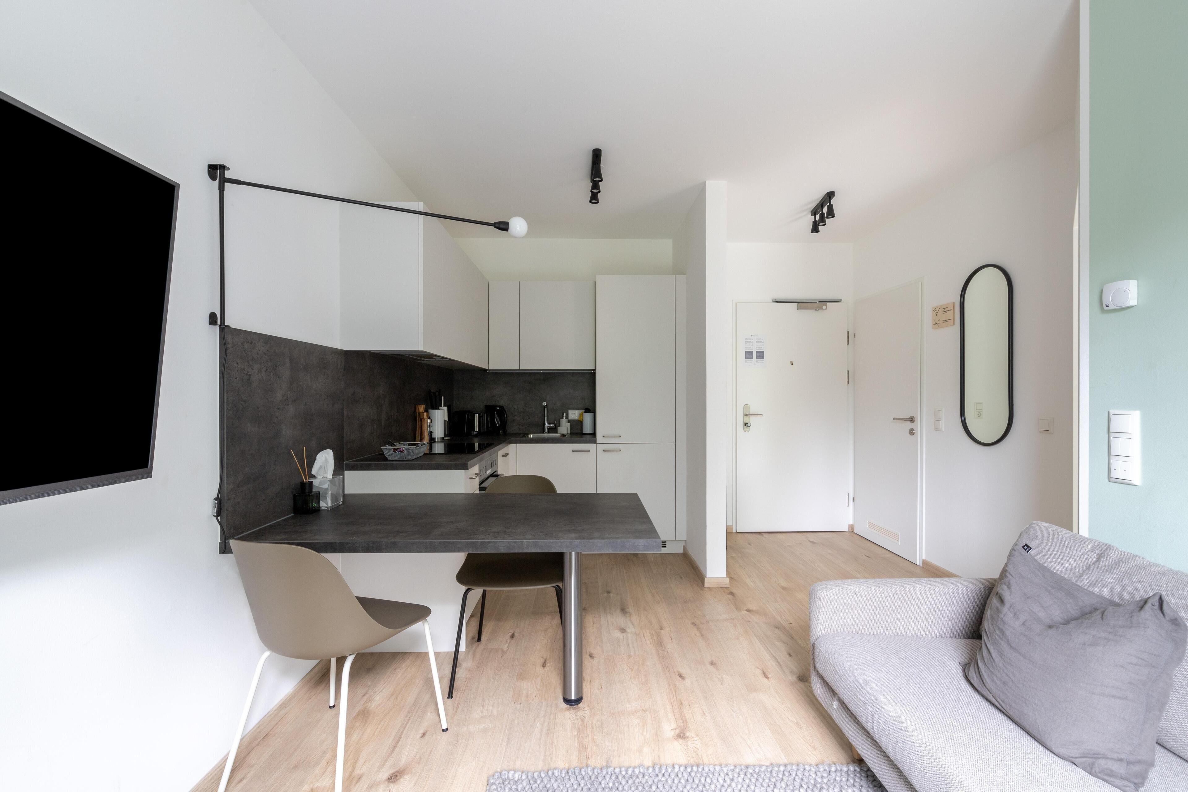 A modern kitchen with a dark countertop, a dining area with a table and chairs, and a living room with a sofa and a TV.
