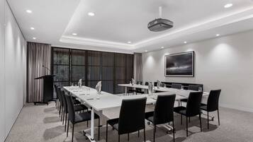 Ein moderner Konferenzraum mit einem langen Tisch, schwarzen Stühlen, einem Projektor und einem großen Fenster mit Vorhängen.