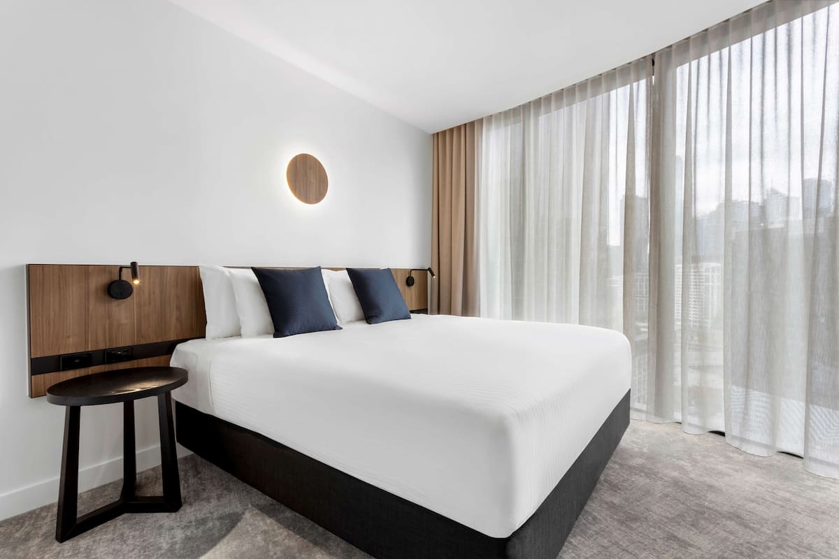 Une chambre d’hôtel moderne avec un grand lit, une applique murale ronde, une petite table d’appoint et des rideaux légers.