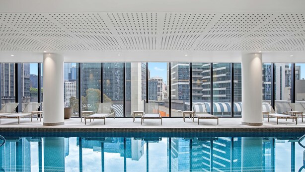 A modern indoor pool with clear glass walls, lounge chairs, and a city view. - Adina Apartment Hotel Melbourne Southbank (Southbank)