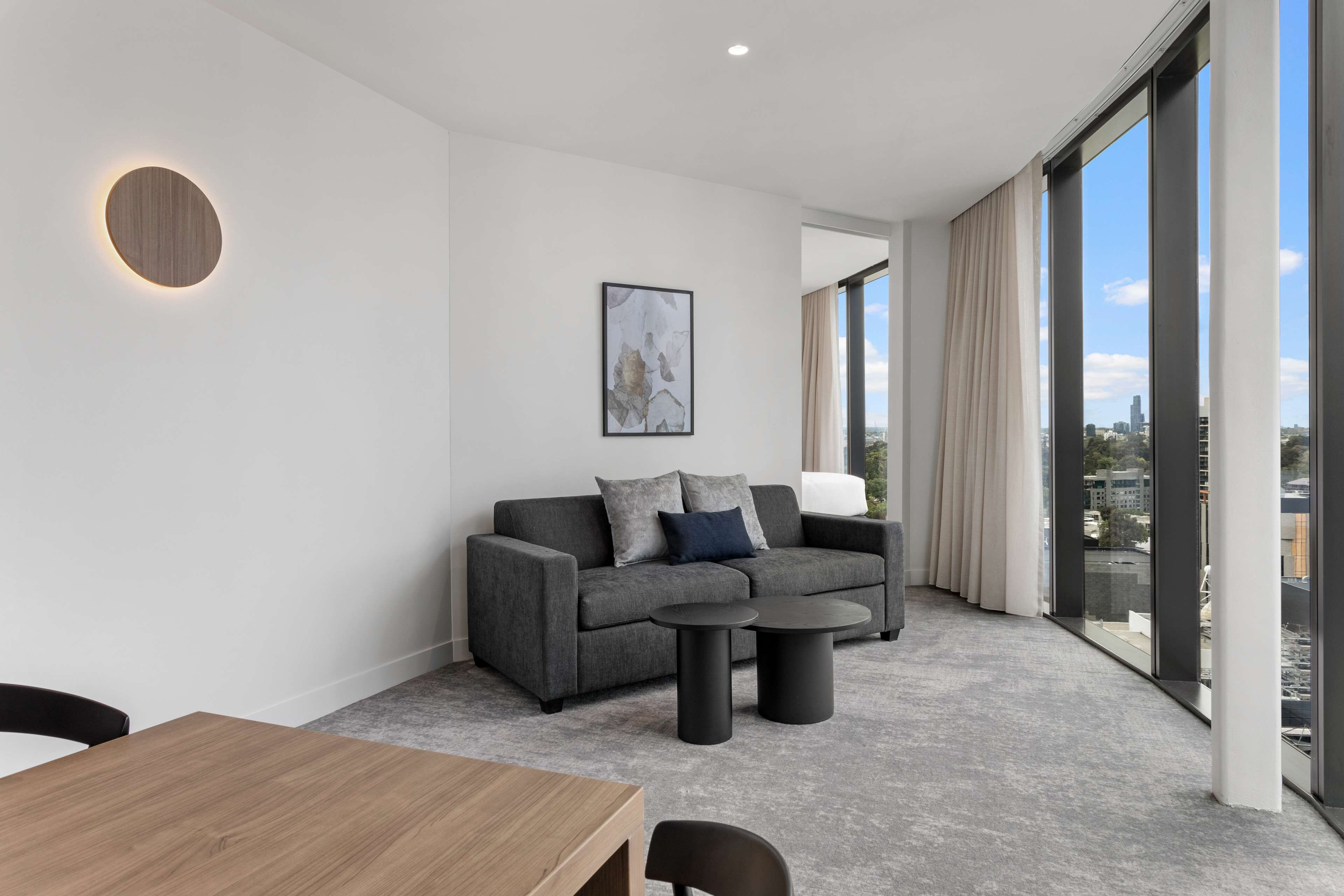 A modern hotel room with a grey sofa, a round table, and a large window offering a city view.
