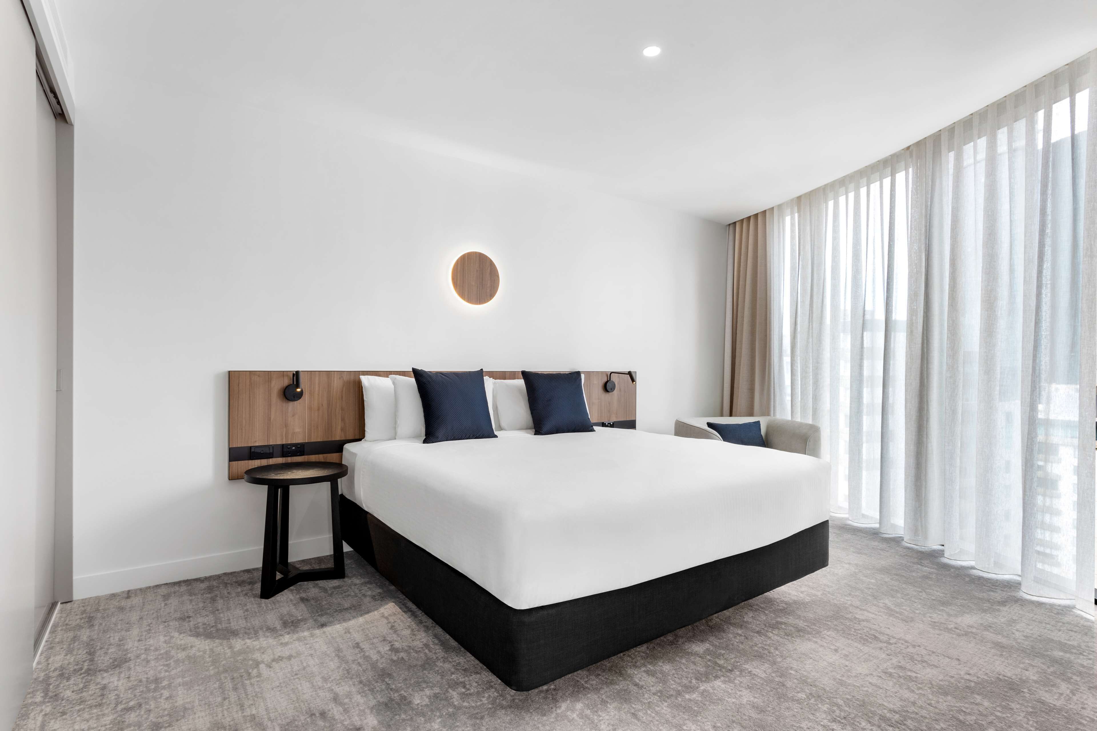 A modern hotel room with a large bed, a round wall light, a small side table, and sheer curtains.