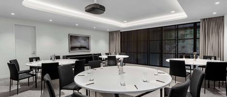 Une salle de conférence moderne, dotée de tables rondes, de chaises noires et d’un grand écran.