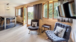 A spacious room with a dining area, two seating options, a television, and large windows offering a view of the outdoors.