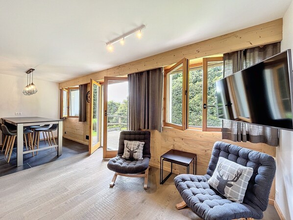 A spacious room with a dining area, two seating options, a television, and large windows offering a view of the outdoors.