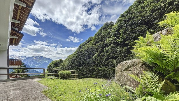 A paved path leading to a building with a balcony overlooking a mountainous landscape.