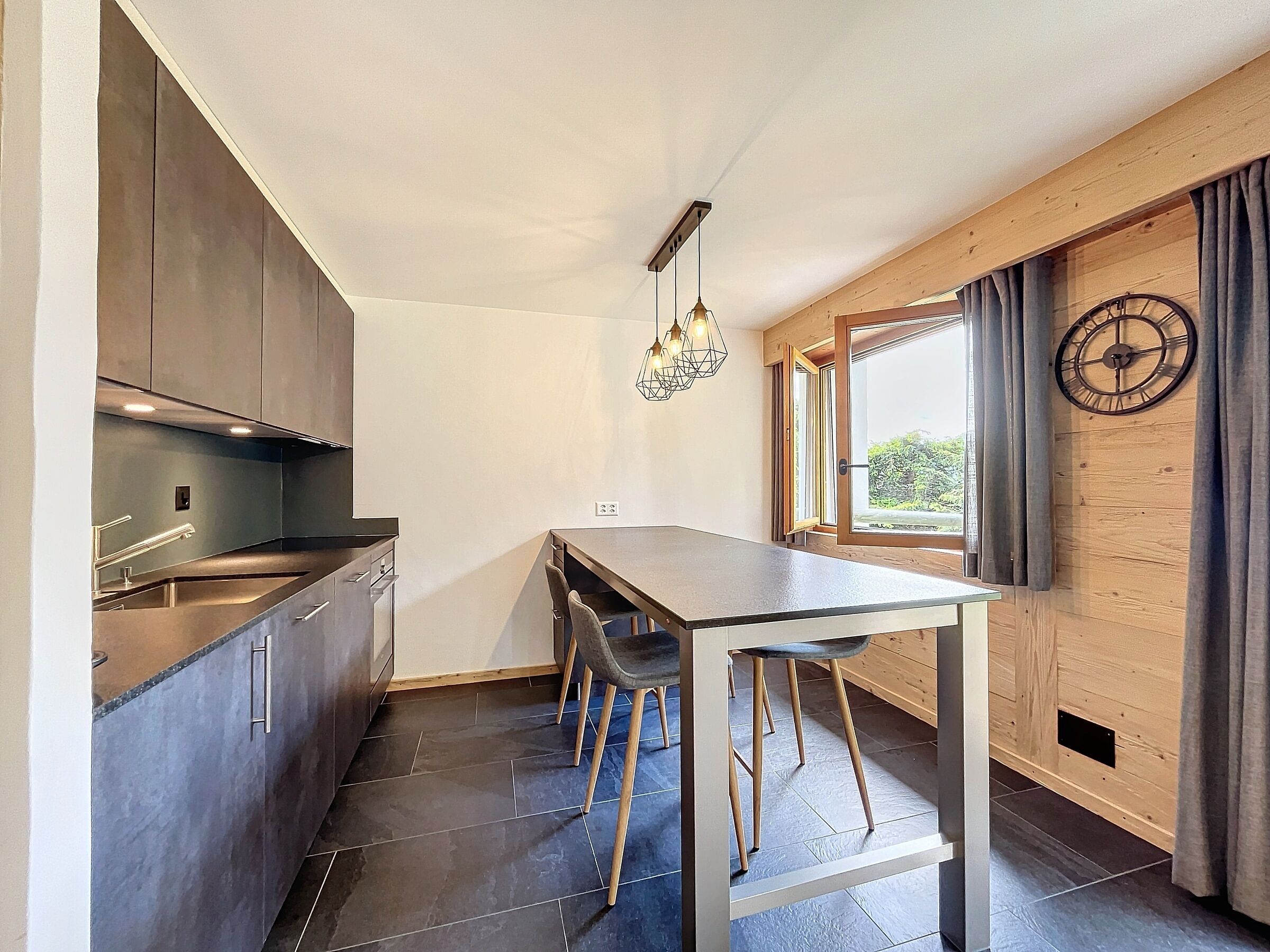 Une cuisine moderne avec des meubles de couleur sombre, un îlot central doté de tabourets, un luminaire suspendu et une fenêtre agrémentée de rideaux.