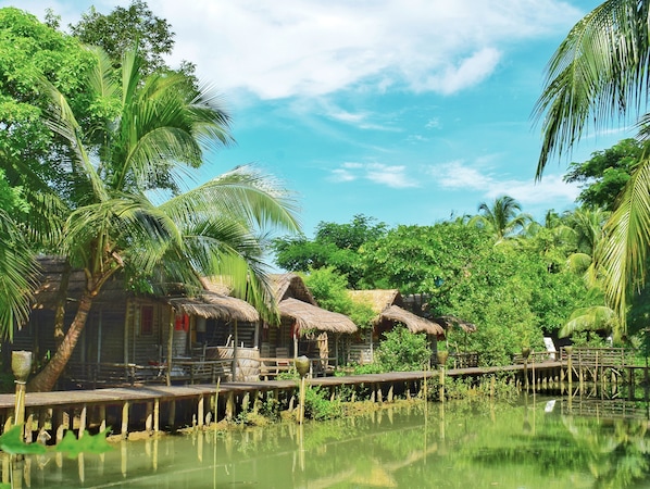 A traditional wooden house with thatched roofs, surrounded by lush greenery and a calm water body.