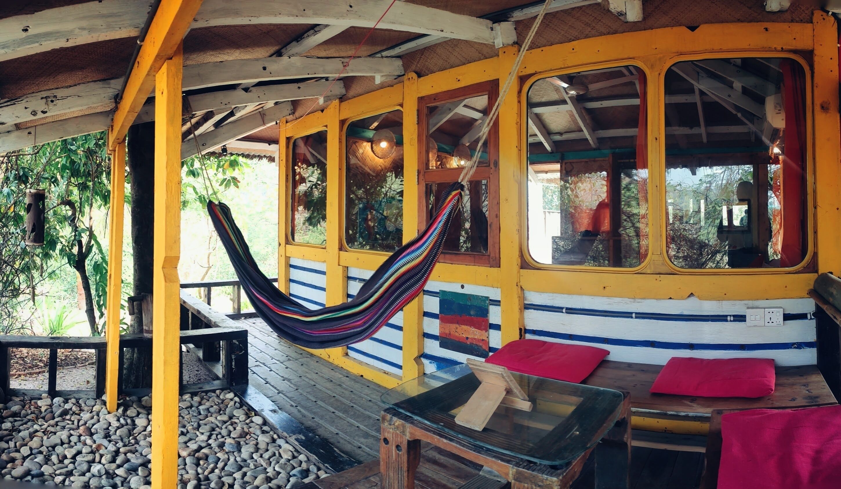 A hammock hanging between yellow and blue walls, a glass table, and red cushions on a wooden bench.