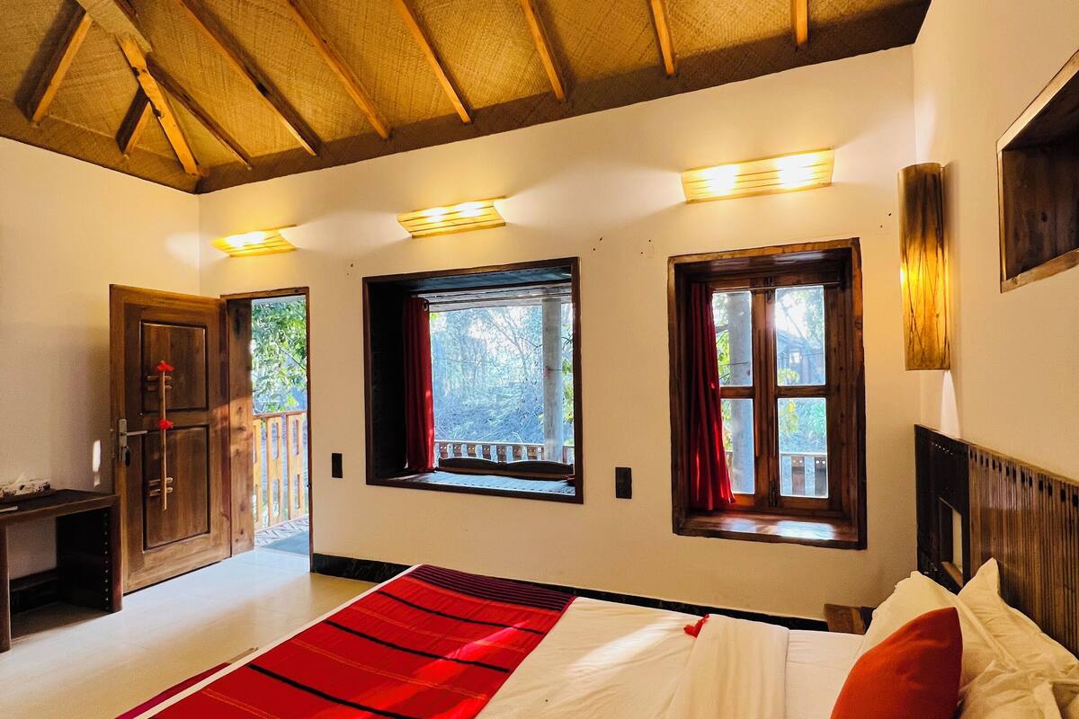 A bedroom with a large bed, wooden ceiling beams, and two windows with curtains.