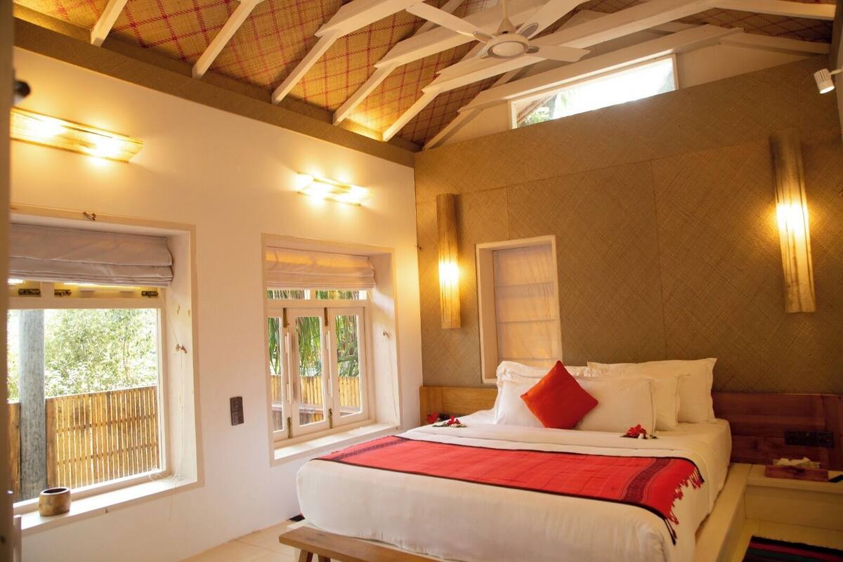 A bedroom with a large bed, two windows, a ceiling fan, and a wooden bench.