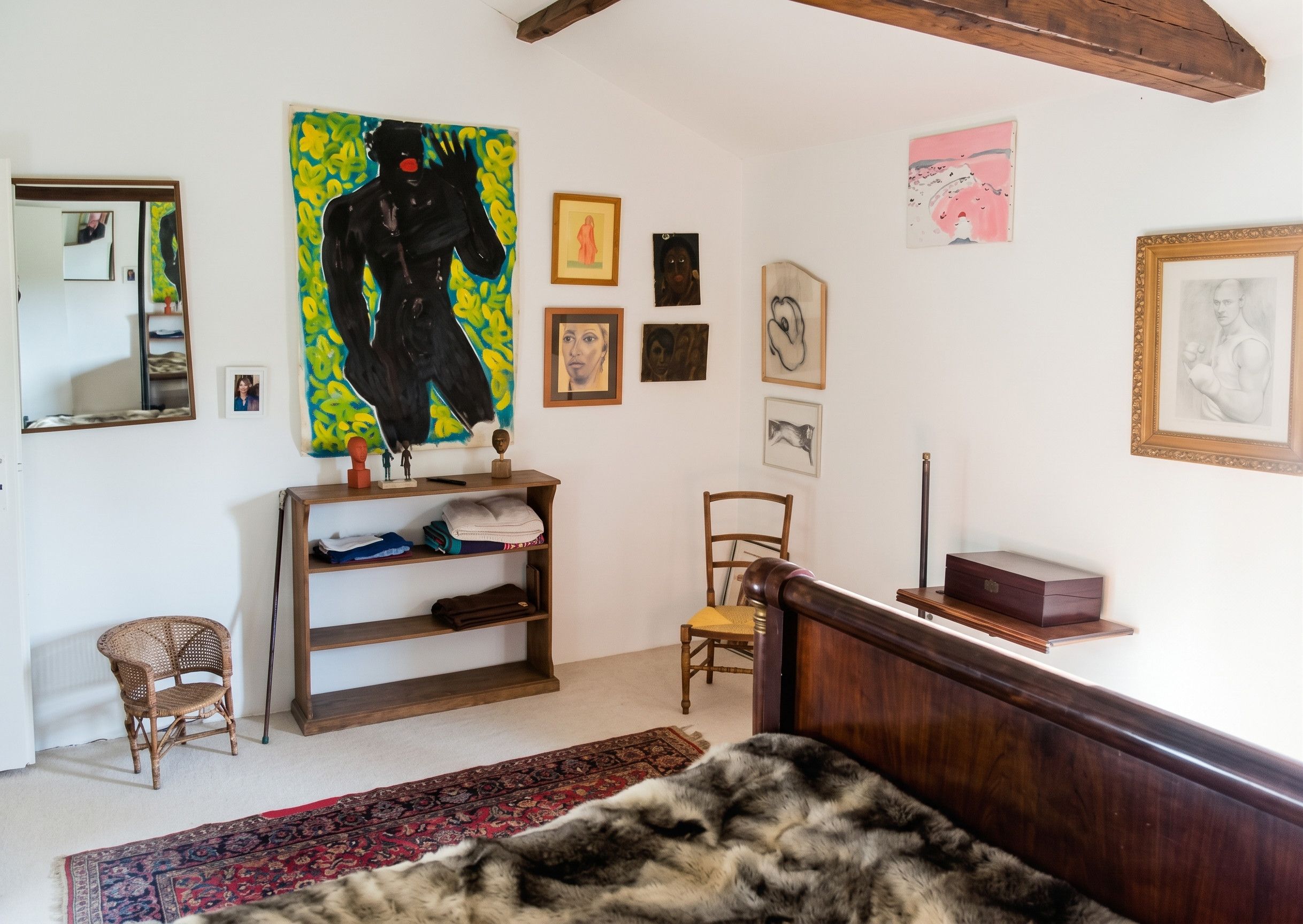 A room with a bed, a wooden headboard, a mirror, a shelf with folded clothes, a chair, and various framed artworks on the wall.