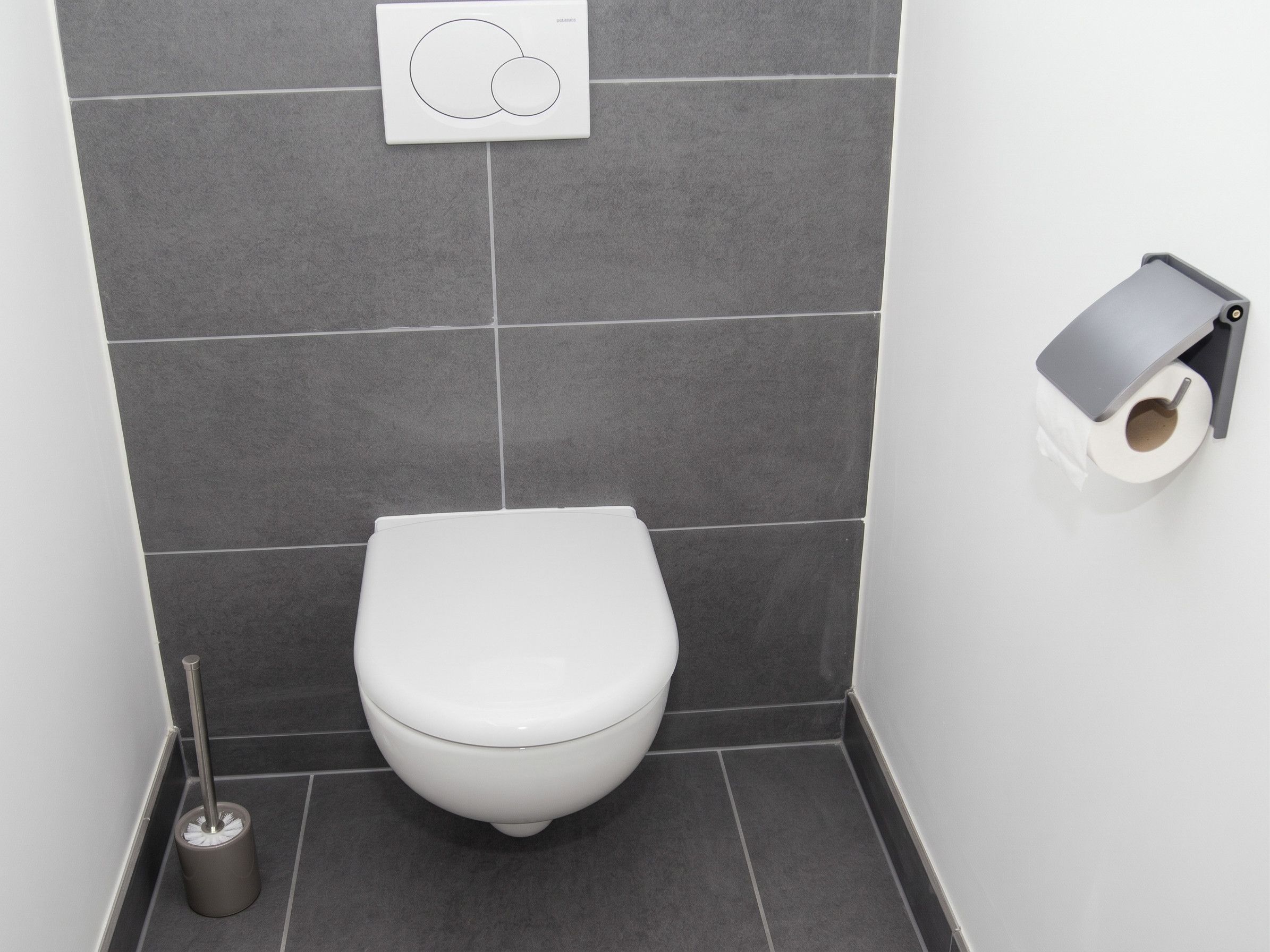 Un baño pequeño y moderno con inodoro blanco, dispensador de papel higiénico y escoba para el piso.