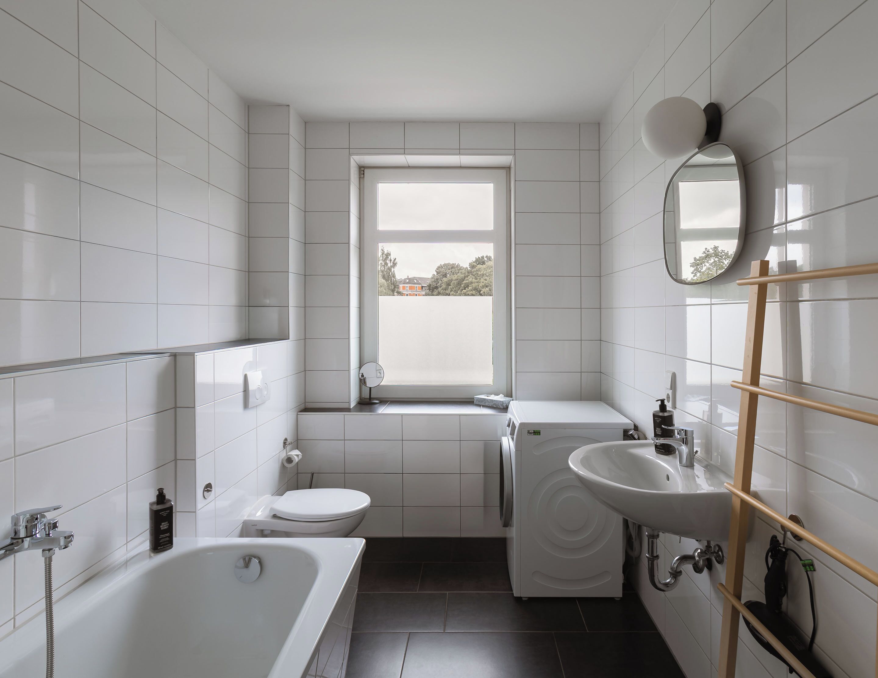 A modern bathroom with a bathtub, toilet, and sink, featuring white tiles and a window.