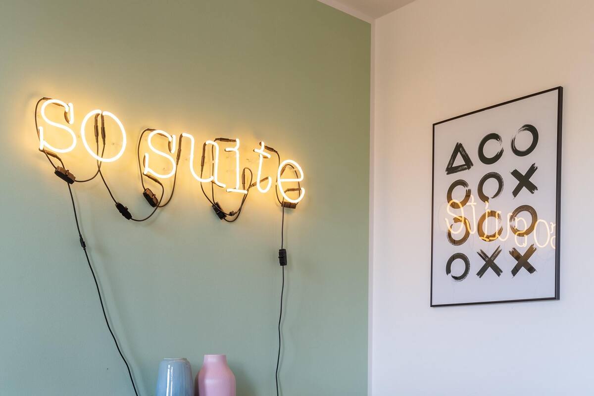 A dining area with a table and four chairs, a neon sign reading "SO SUITE", and a framed abstract artwork on the wall.