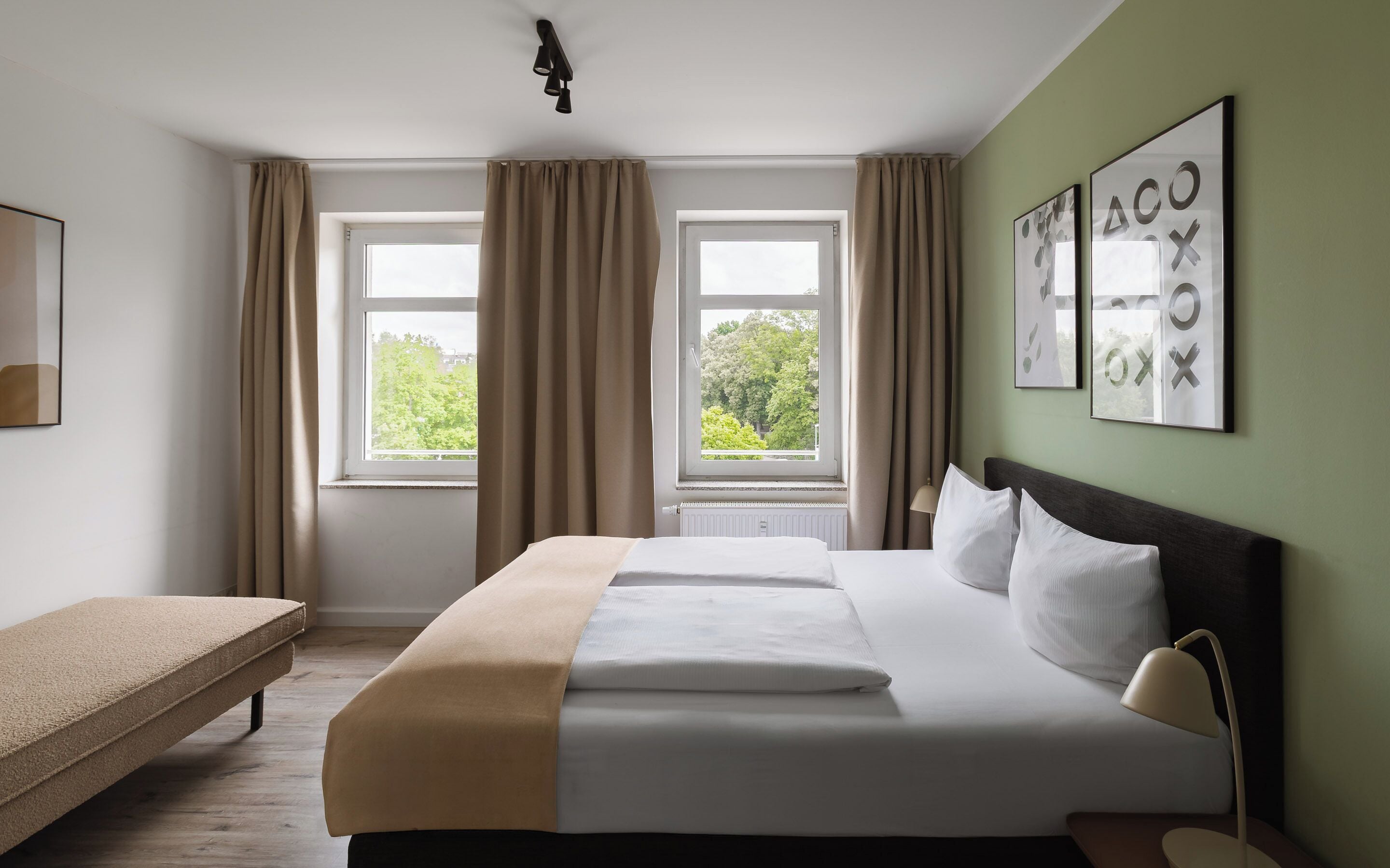 Ein Hotelzimmer mit einem Bett, einer Sitzbank, zwei mit Vorhängen versehenen Fenstern und Bildern an der Wand.