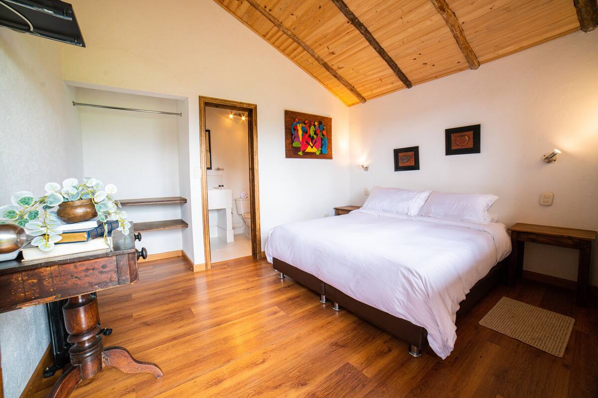 Een slaapkamer met een houten plafond, een bed met wit beddengoed, een houten bureau met een vaas en een schilderij aan de muur.