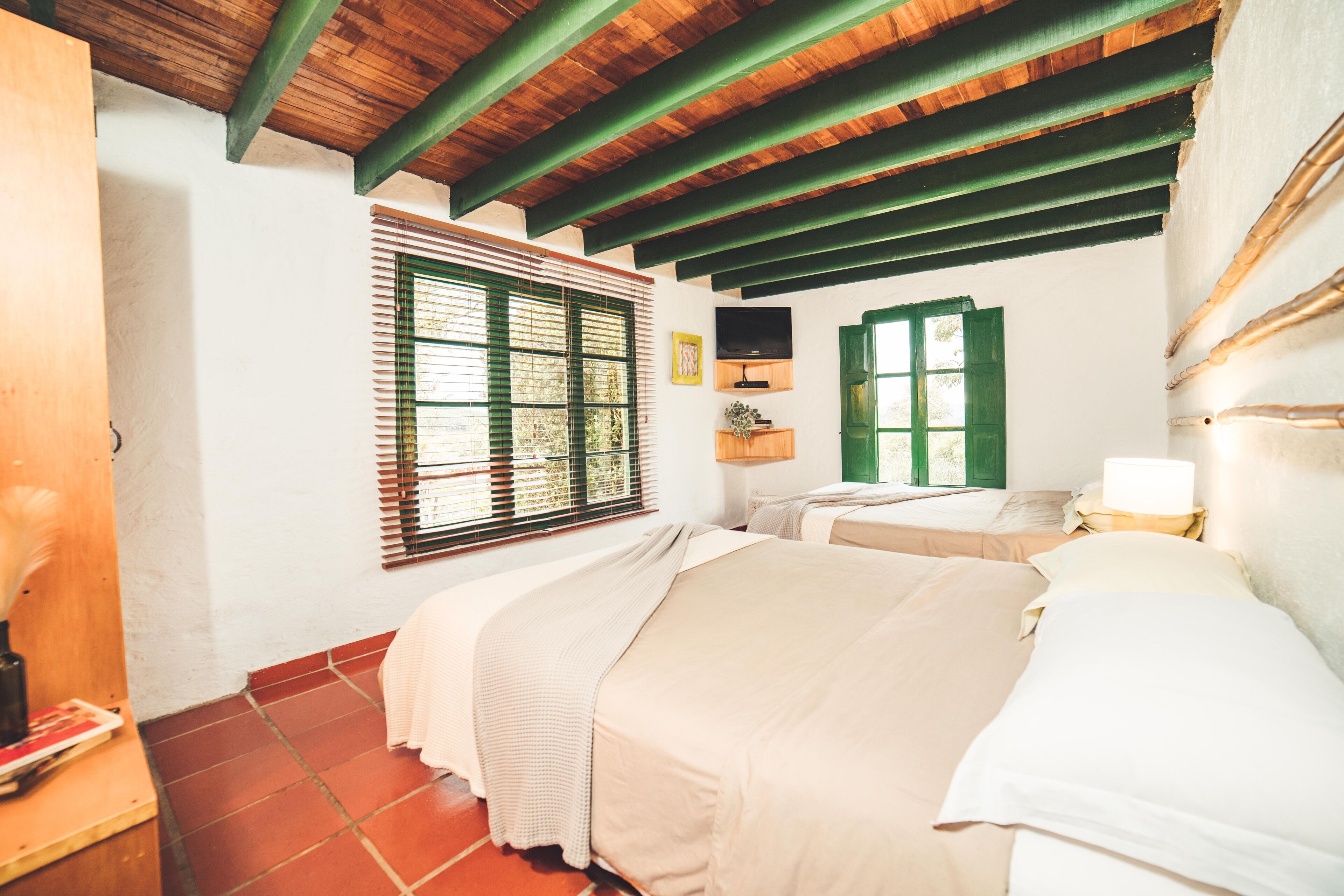 A bedroom with two beds, wooden beams on the ceiling, a window with blinds, and a television on the wall.