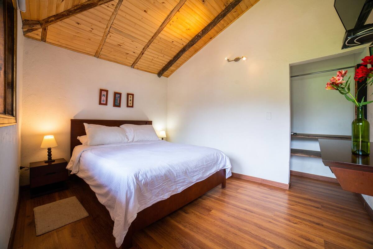 Een slaapkamer met een houten plafond, een bed met witte beddengoed, een nachtkastje met een lamp en een bloemenvaas op een dressoir.