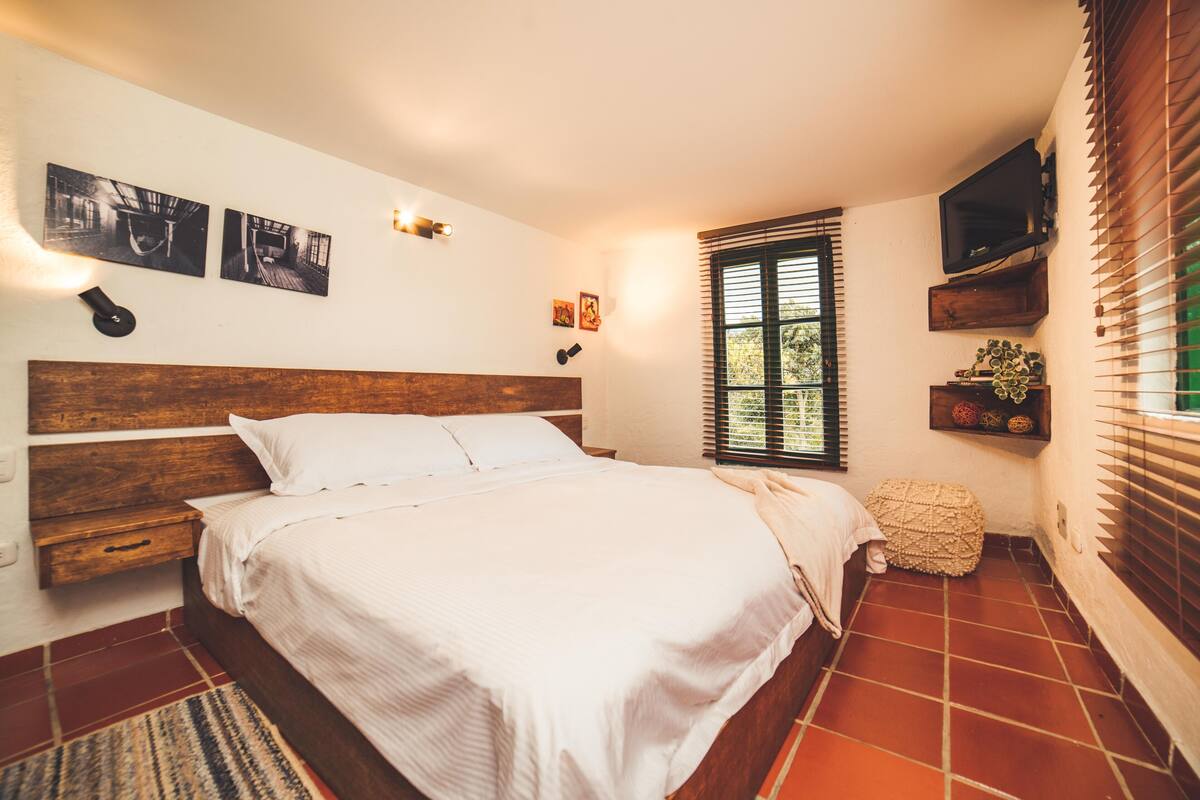 Een slaapkamer met een houten hoofdbord, een bed met witte lakens, een klein nachtkastje, een tv aan de muur en een raam met jaloezieën.