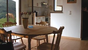 A dining area with a round wooden table and chairs, a vase with flowers, and a view of a cow grazing outside.