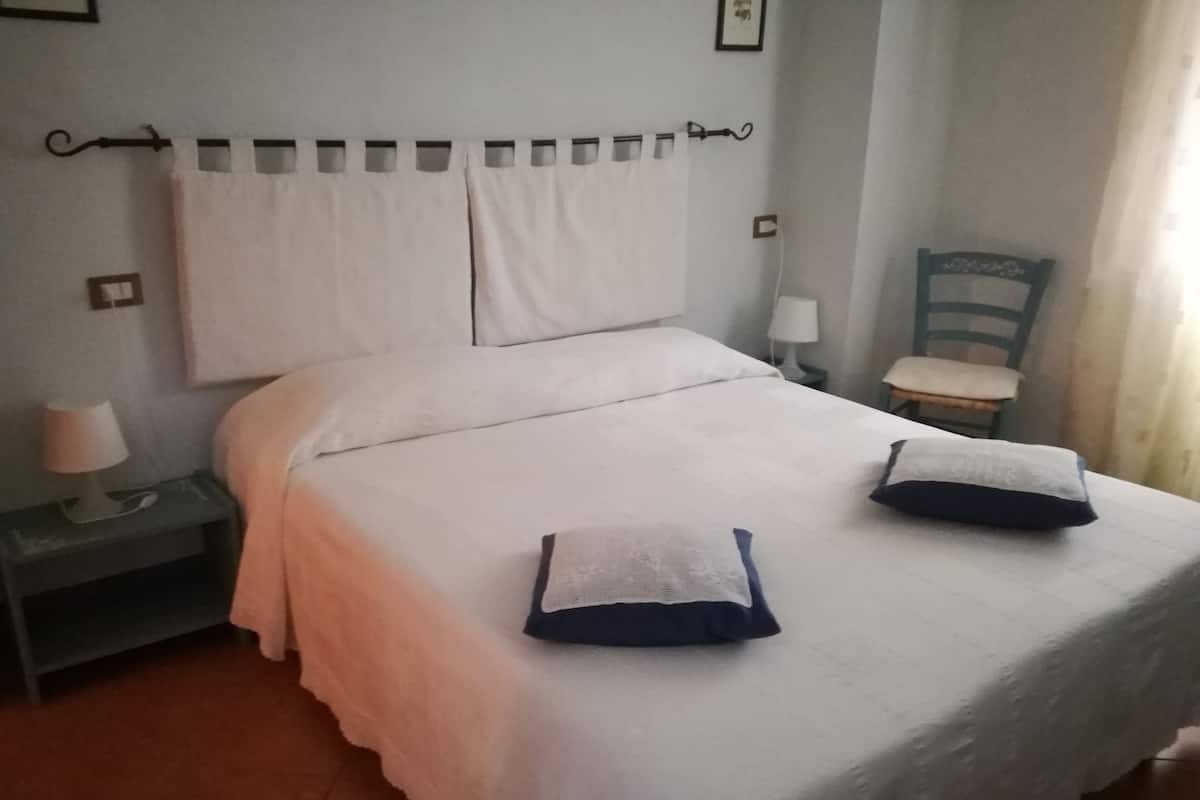 Cama bem arrumada com almofadas brancas e azuis, mesa de cabeceira com abajur, e uma cadeira em um quarto simples.