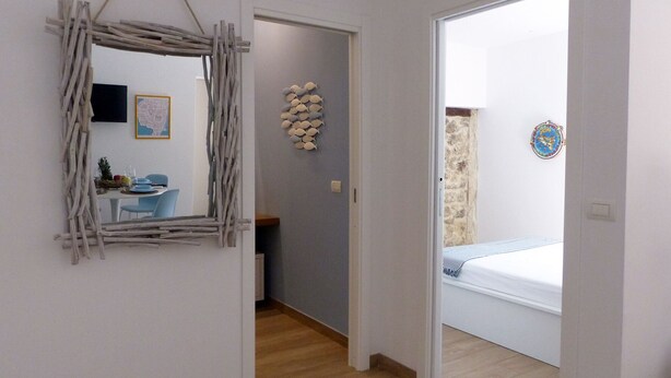Una habitación con piso de madera, una cama con colchón blanco, una mesita con una silla, un espejo montado en la pared y un adorno de peces en la pared.