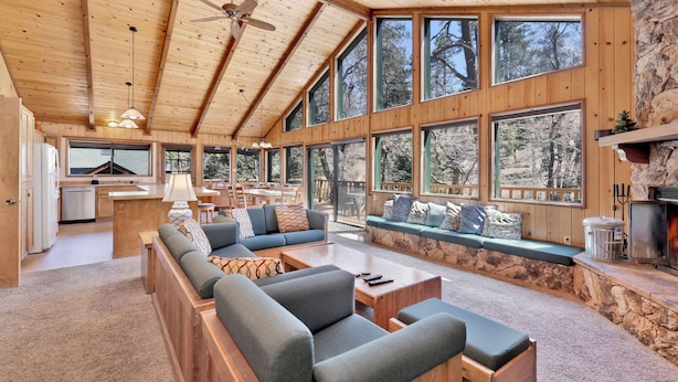 A spacious living area with a long sofa, a coffee table, and a stone fireplace. Large windows provide views of the surrounding forest. - Wodehouse #1057 (Big Bear Lake)