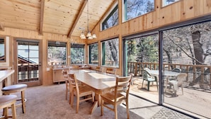 A wooden interior with a large dining table, chairs, and a view of the outdoors through large windows. - Wodehouse #1057 (Big Bear Lake)