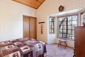 A bedroom with a quilted bedspread, a wooden door, a window with a view of trees, and a wall clock.