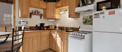Una cocina con armarios de madera, estufa, refrigerador y un comedor con una silla.