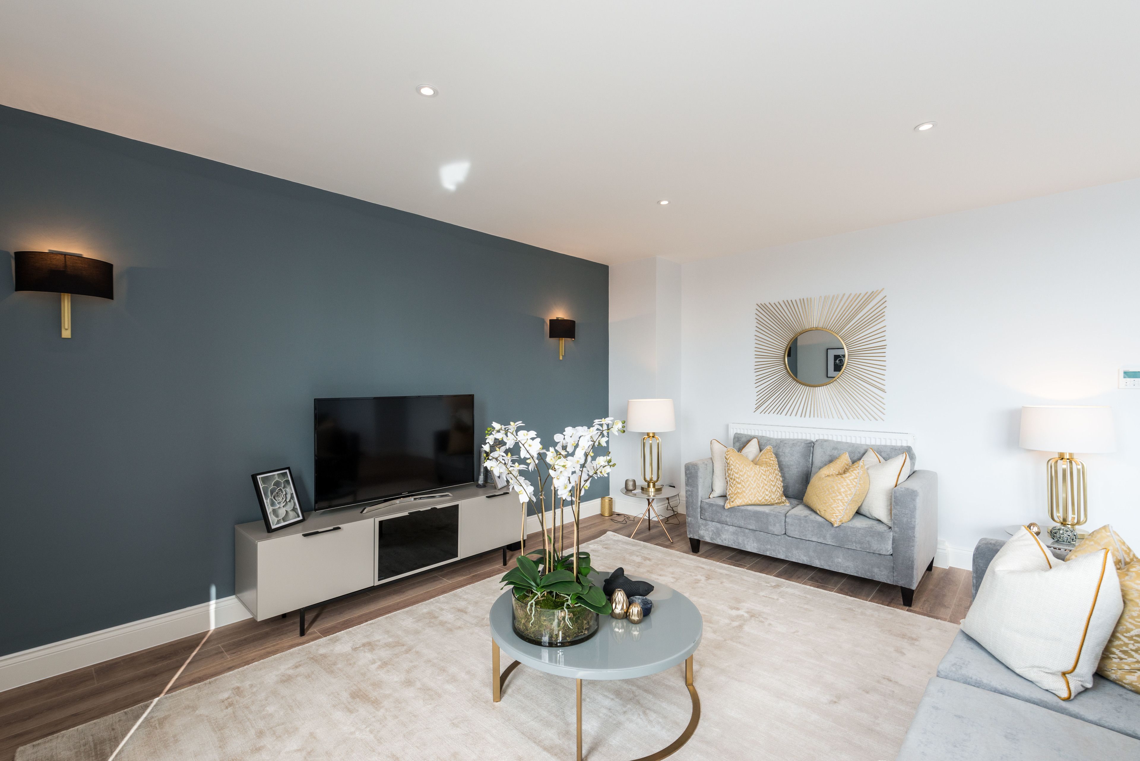 A modern living room with a flat-screen TV, a coffee table with decorative items, a grey sofa with yellow cushions, and a wall-mounted mirror.