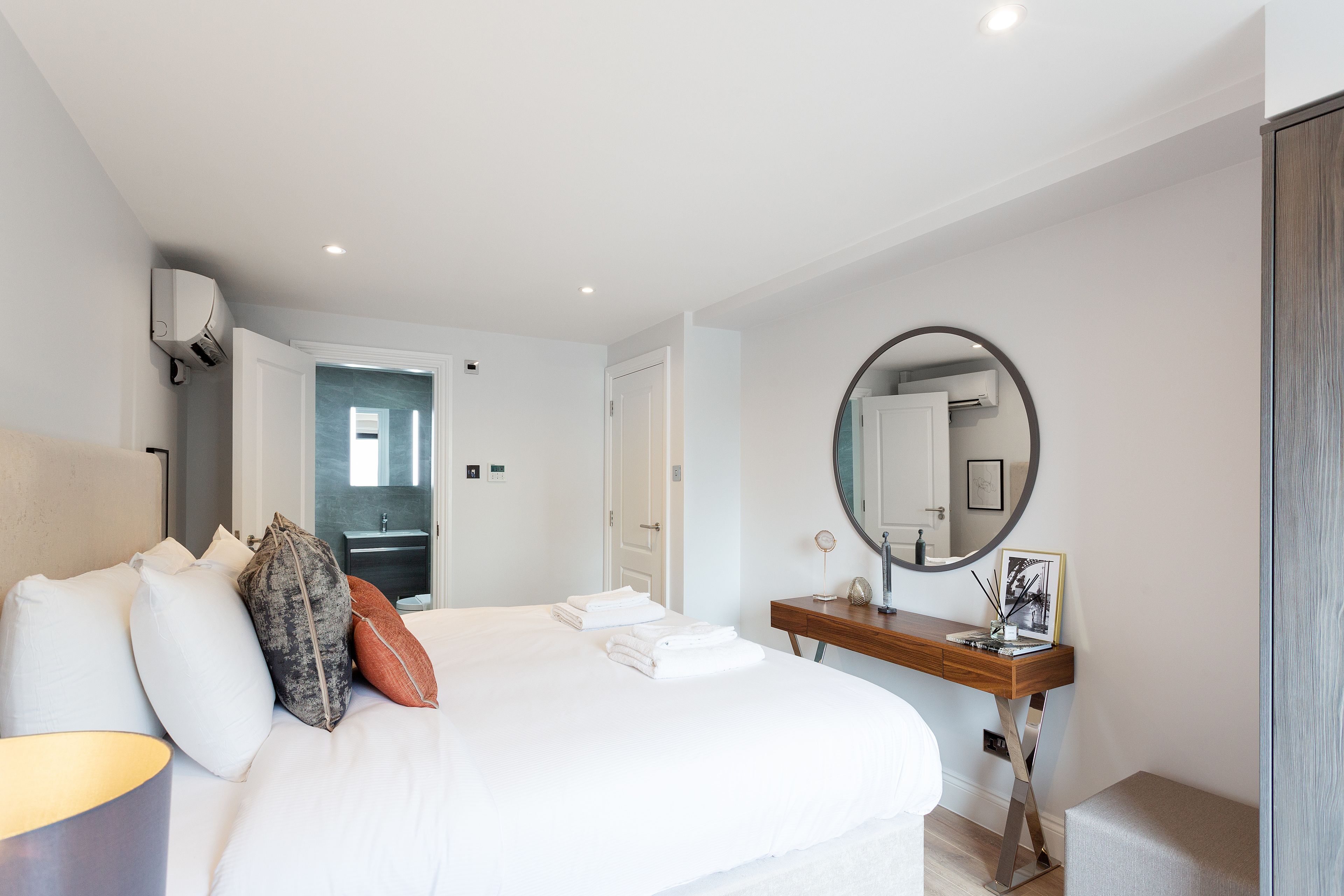 Une chambre d’hôtel moderne avec un grand lit, un bureau en bois, un miroir rond et une salle de bain visible à travers une porte ouverte.