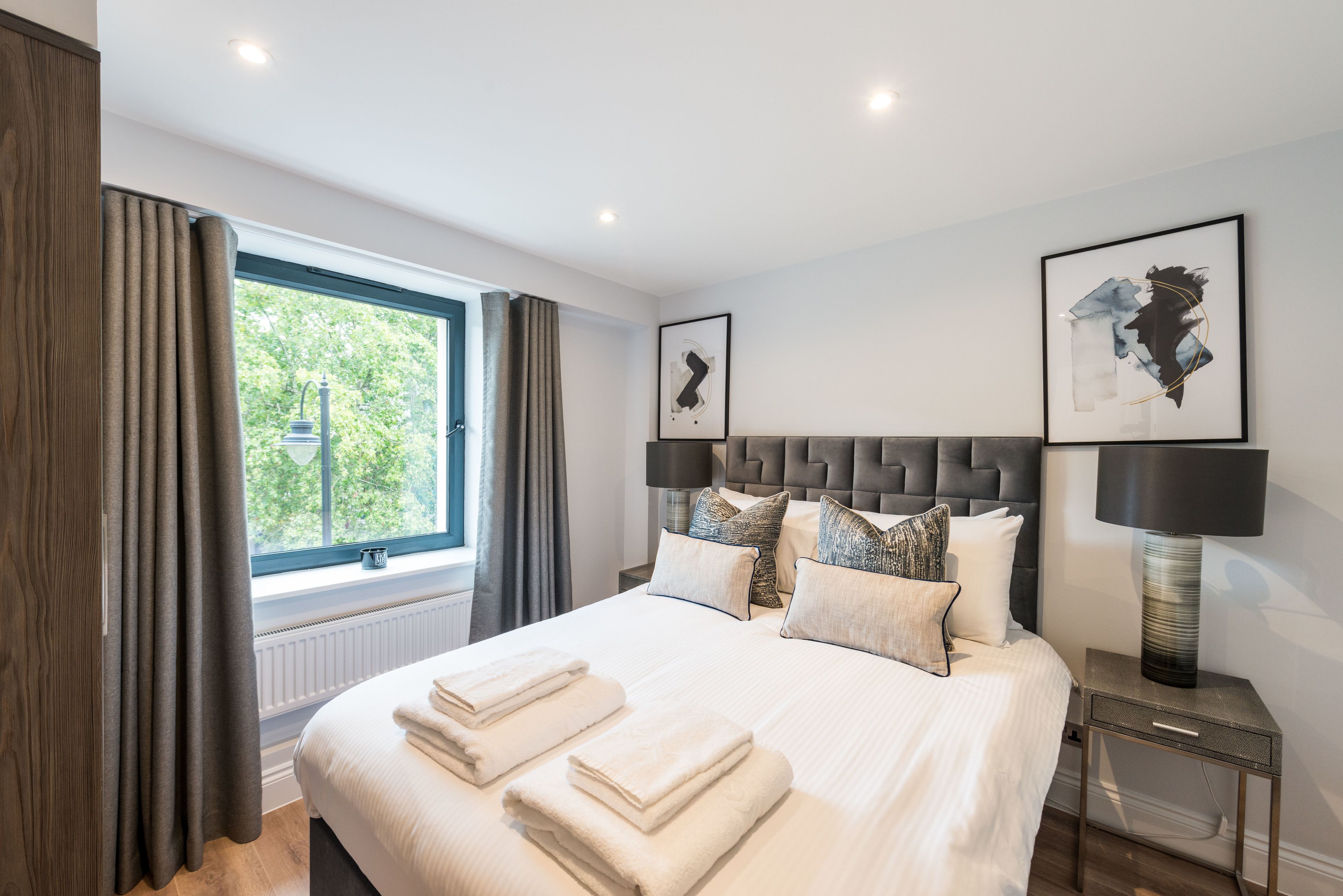 A neatly made bed with white linens and folded towels, a modern headboard, and two framed artworks on the wall.