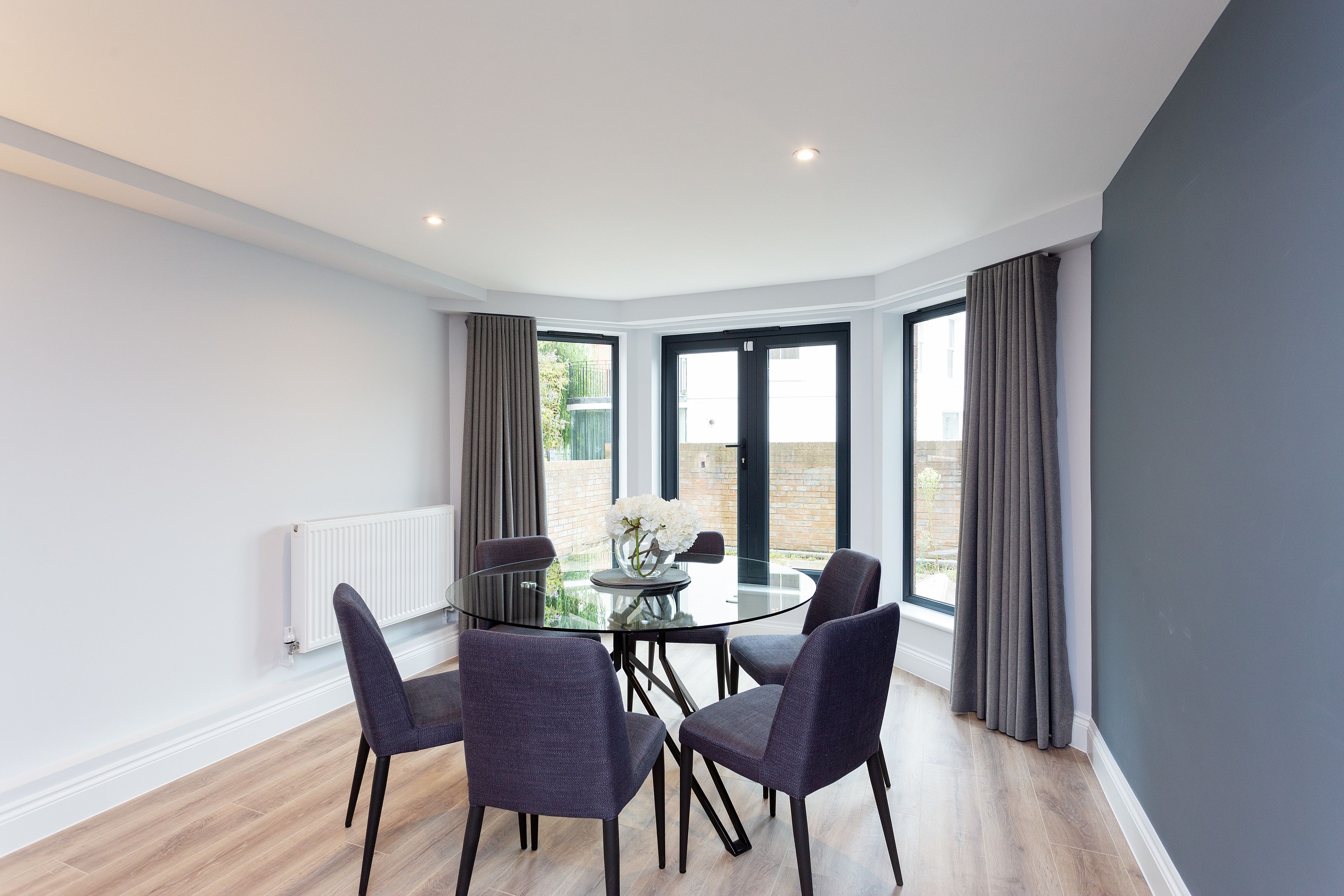 A modern dining area with a glass table and four chairs, large windows with curtains, and a view of greenery outside.