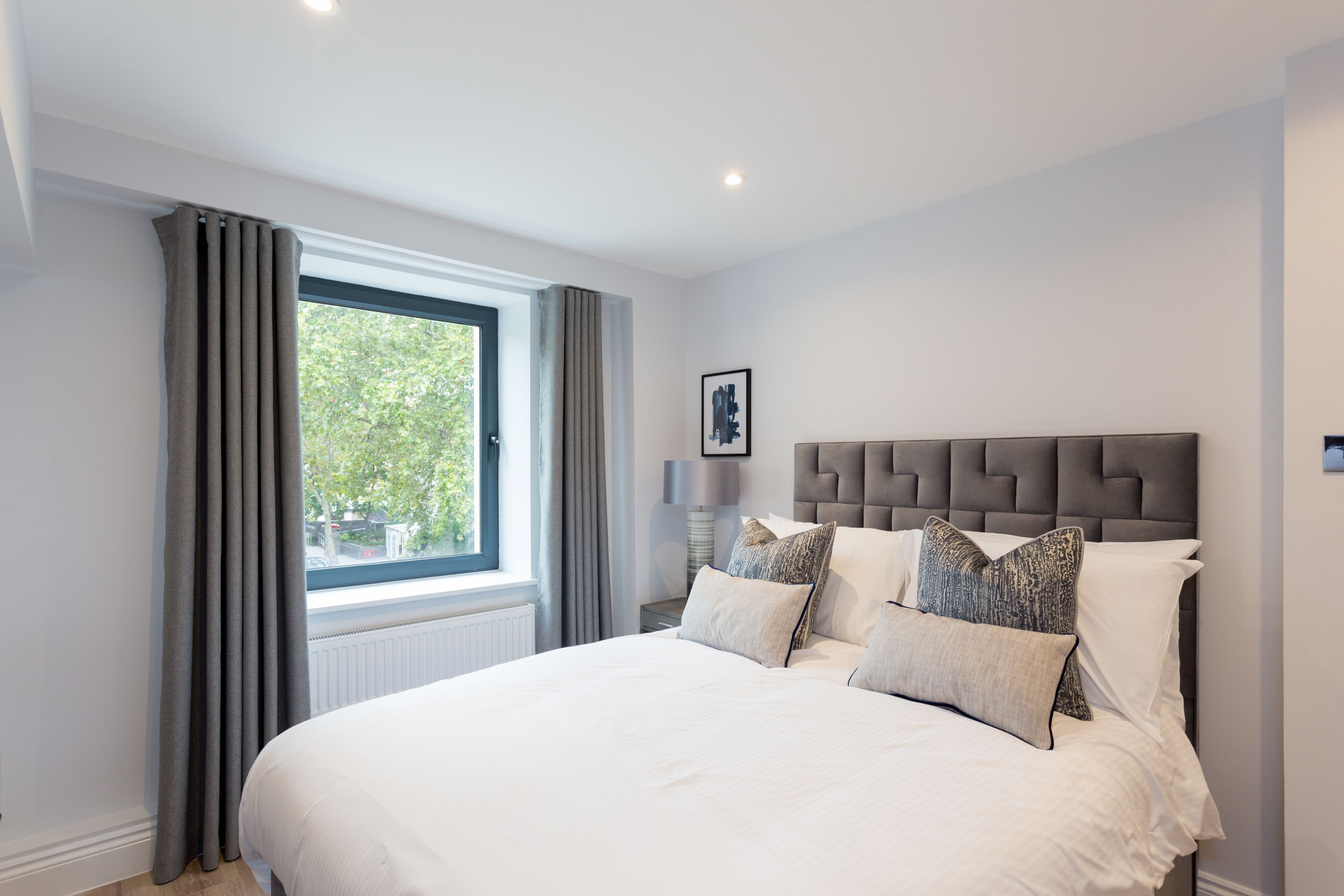 A bedroom with a large bed, grey curtains, a window with a view of greenery, and a modern headboard.