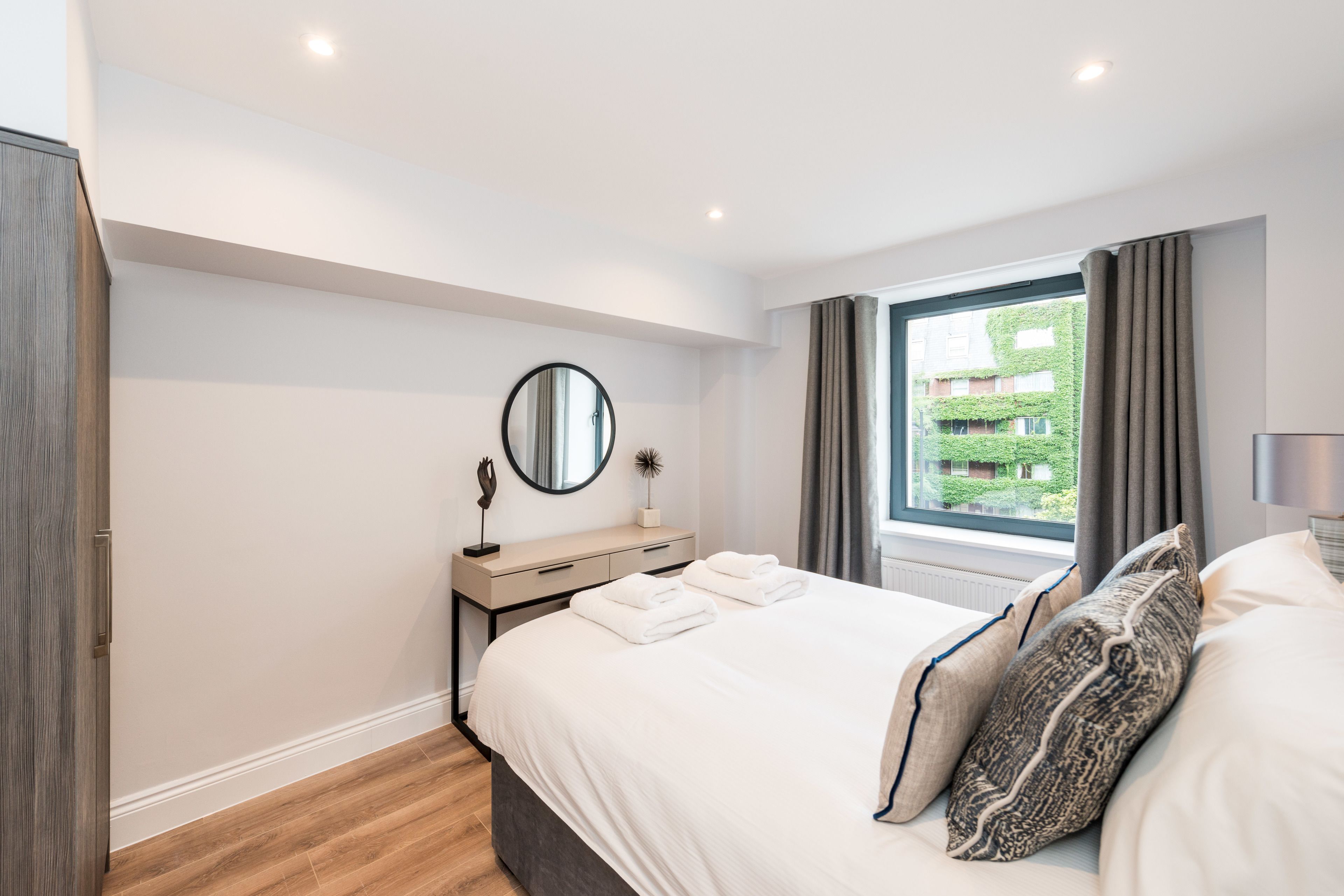 A modern bedroom with a large bed, a bedside table with a mirror, and a window with curtains.