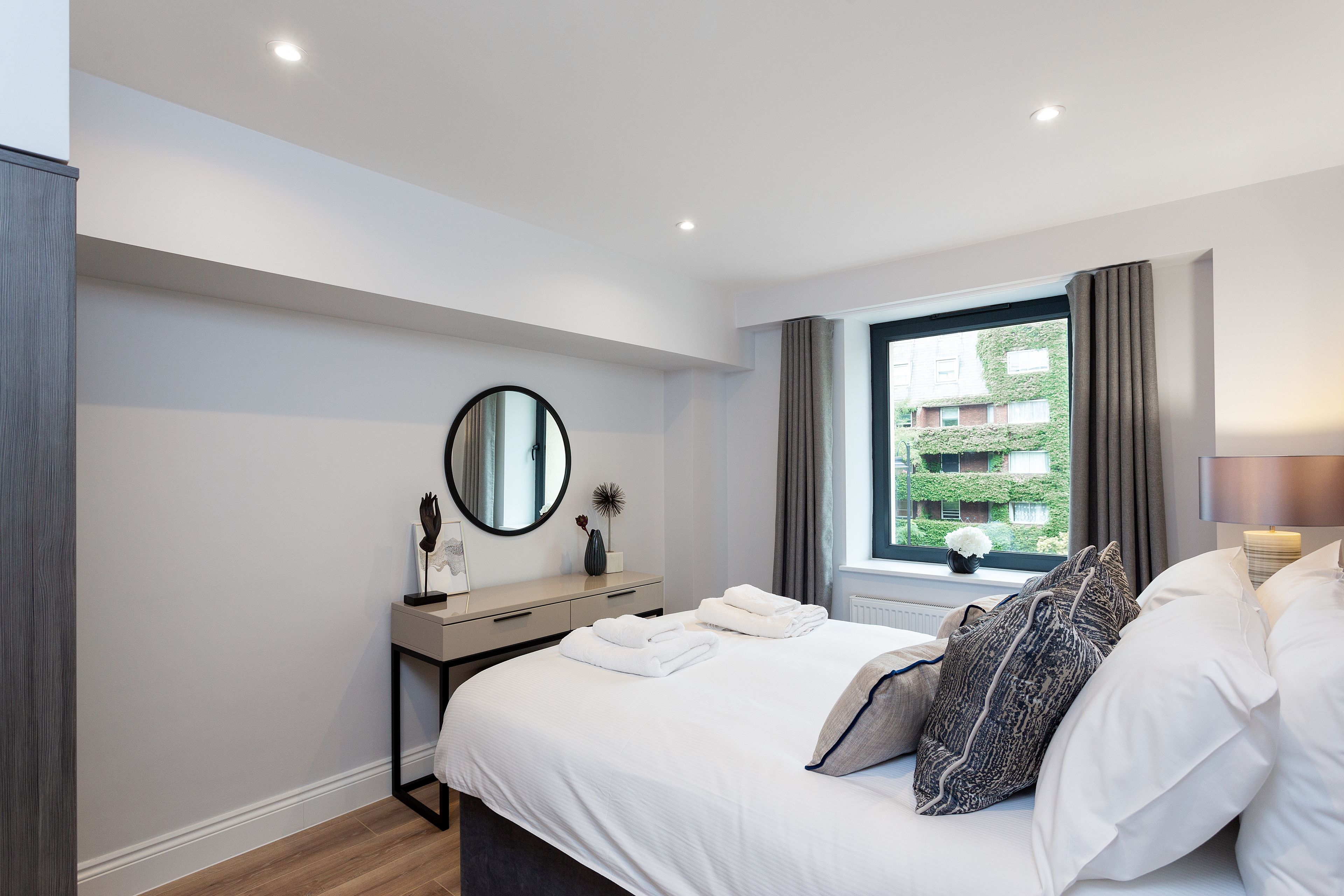 A modern hotel room with a neatly made bed, a bedside table with a lamp, a window with curtains, and a view of greenery outside.