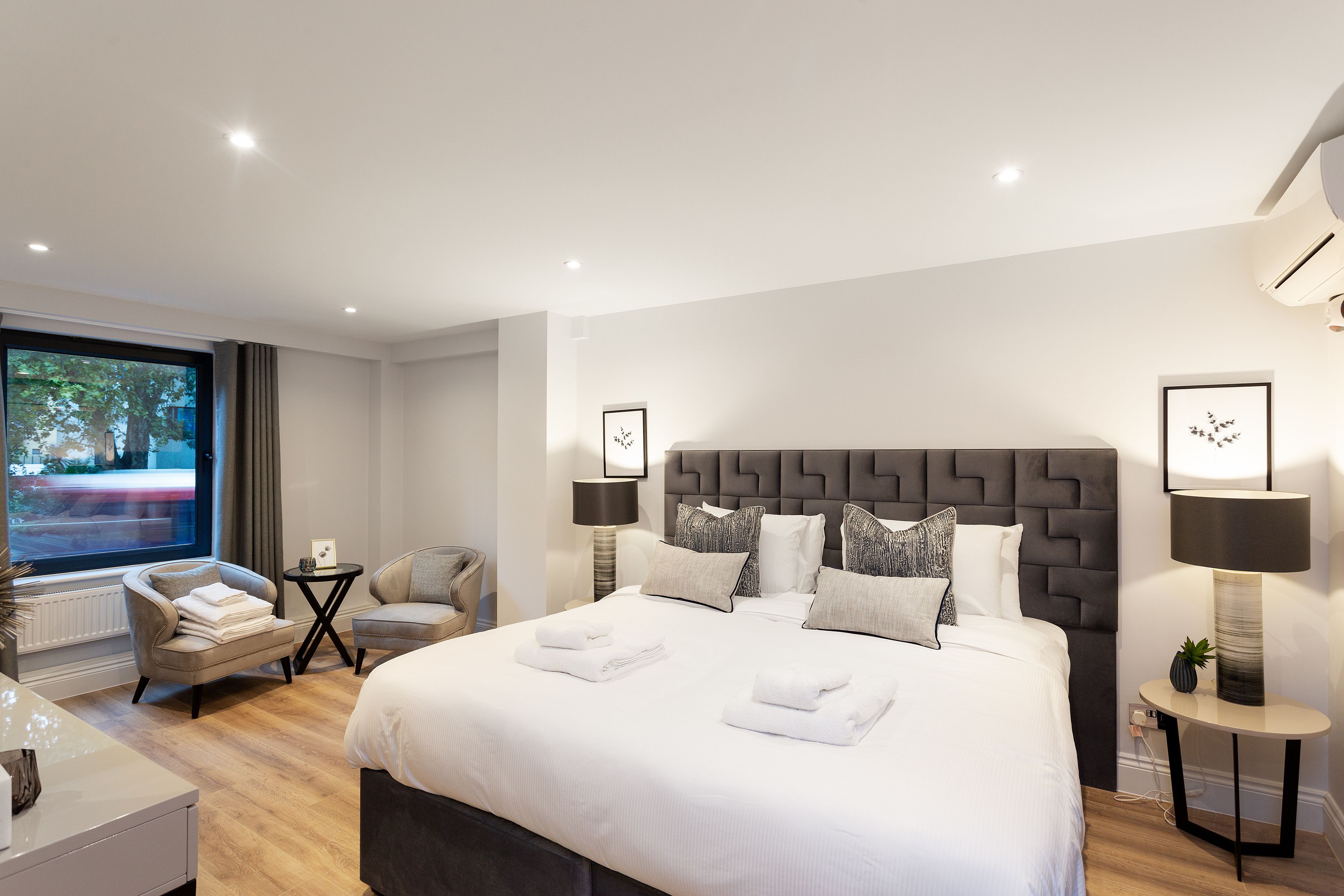 A modern hotel room with a large bed, two armchairs, a small table, and a view of the street through a large window.