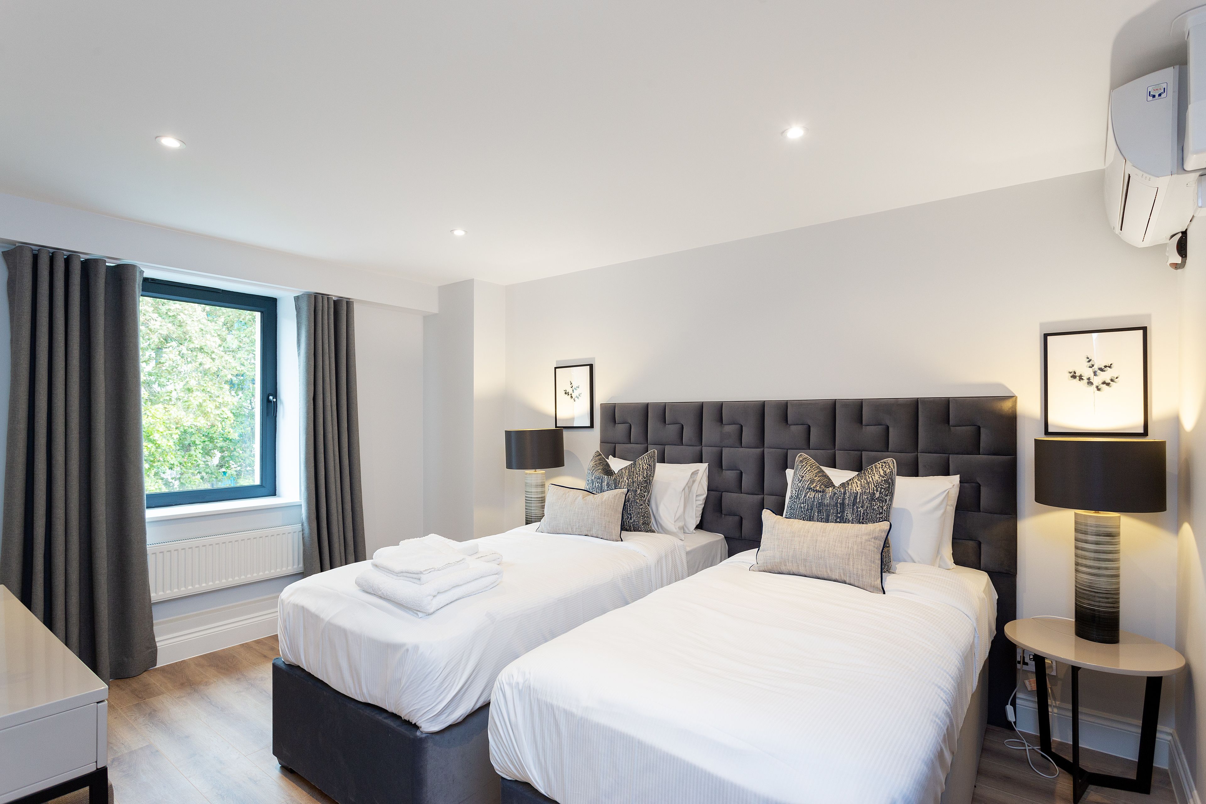 A modern hotel room with two beds, a large window with curtains, a round side table with a lamp, and a wall-mounted air conditioner.