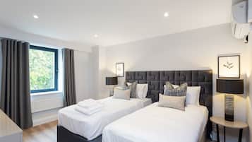 A modern hotel room with two beds, a large window with curtains, a round side table with a lamp, and a wall-mounted air conditioner.
