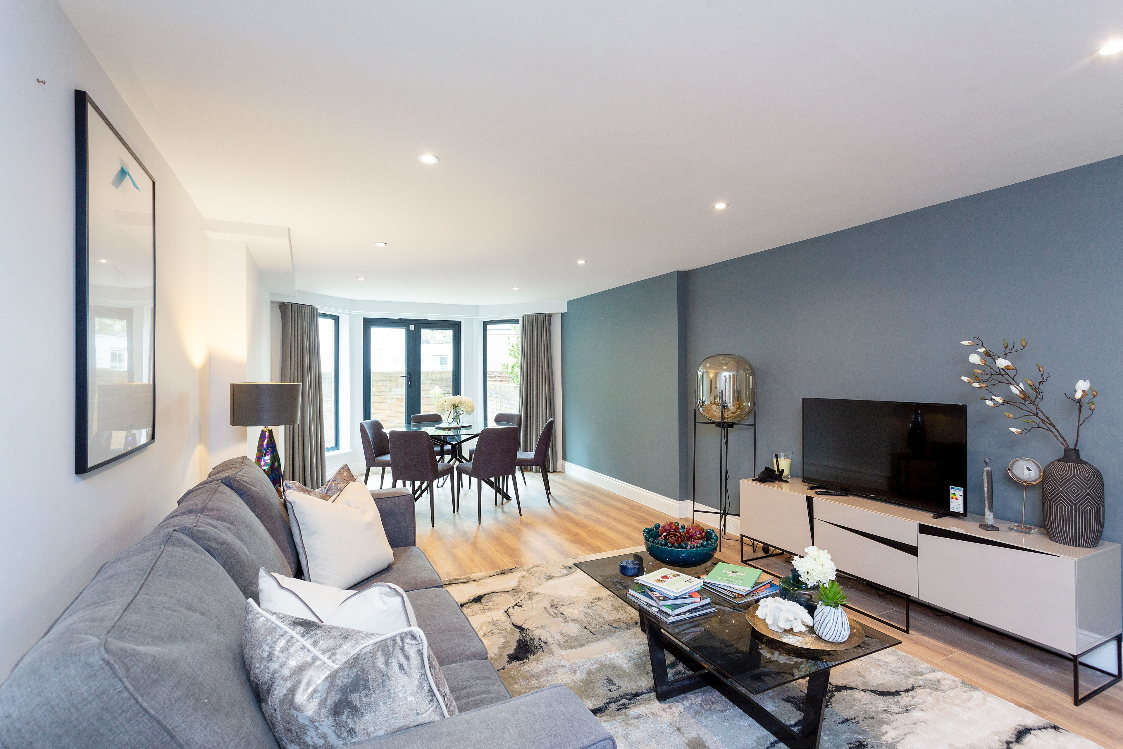 A modern living room with a grey sofa, a coffee table with books and flowers, a TV, and a dining area with chairs and a table.