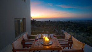 Een houten tafel met kaarsen en wijnglazen buiten, met uitzicht op een mooi landschap bij zonsondergang.