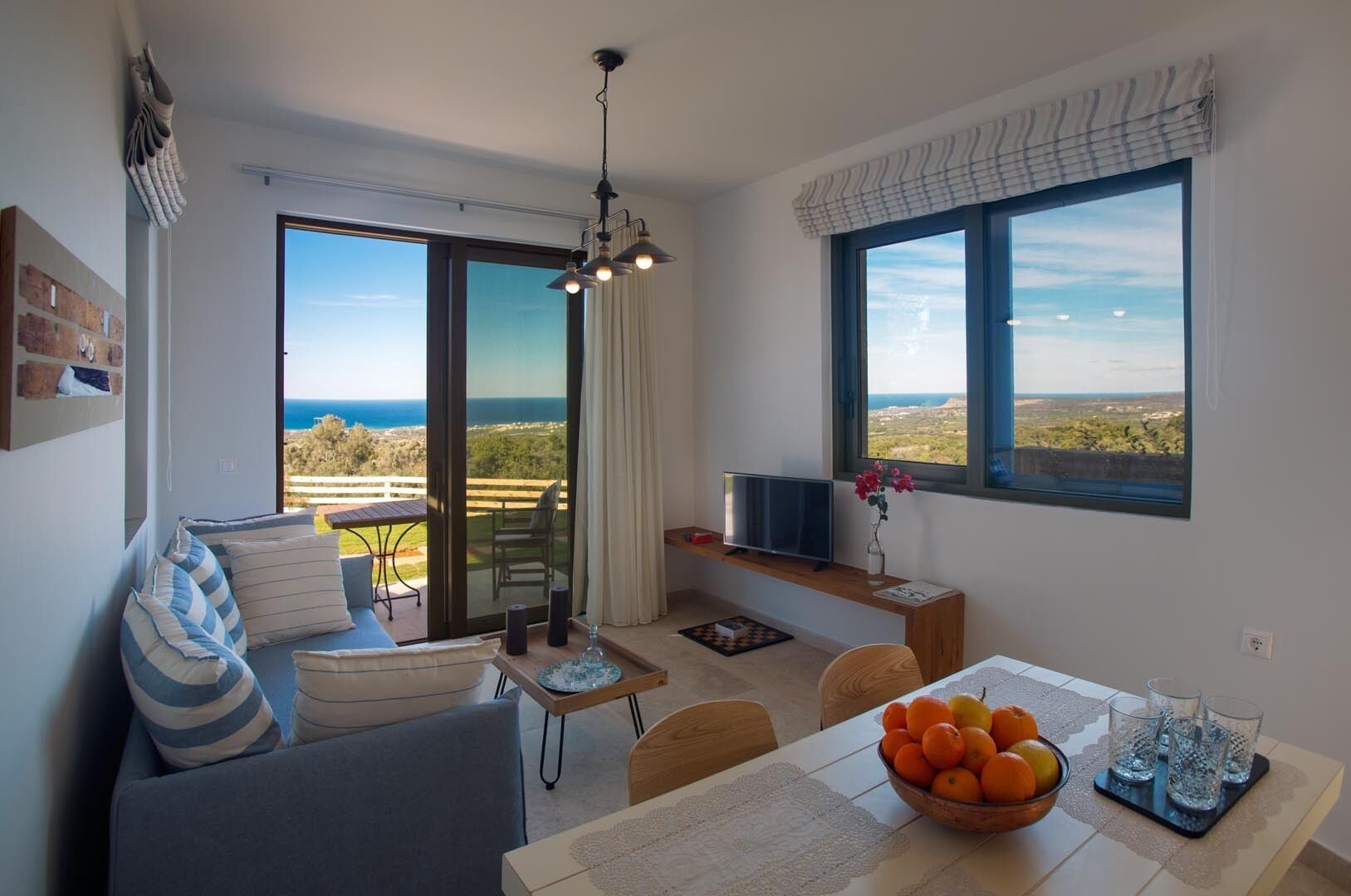 Un salon moderne avec un canapé, une table à manger agrémentée d’un panier de fruits, un téléviseur et une vue sur la mer grâce à de grandes fenêtres.