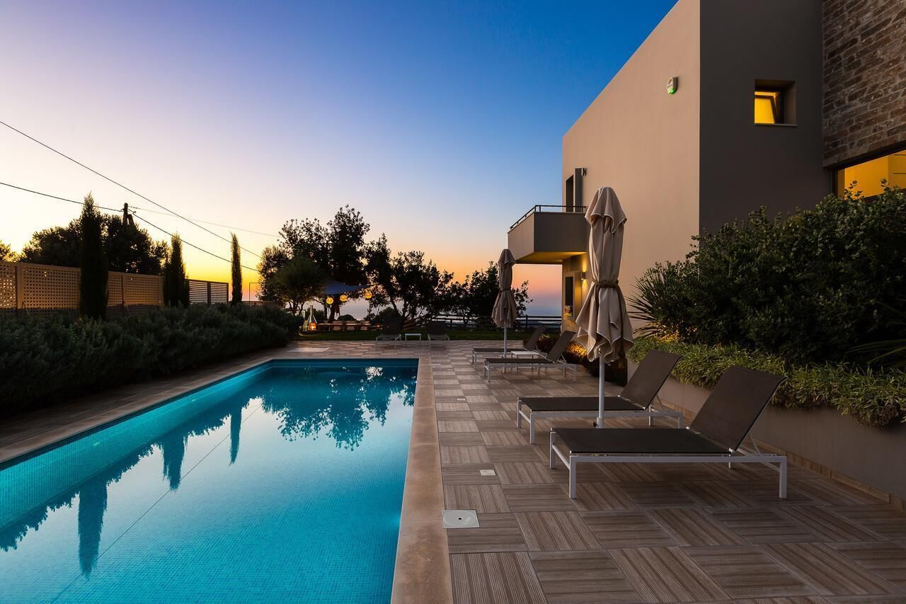 A swimming pool with lounge chairs and umbrellas, surrounded by a modern building with a balcony.