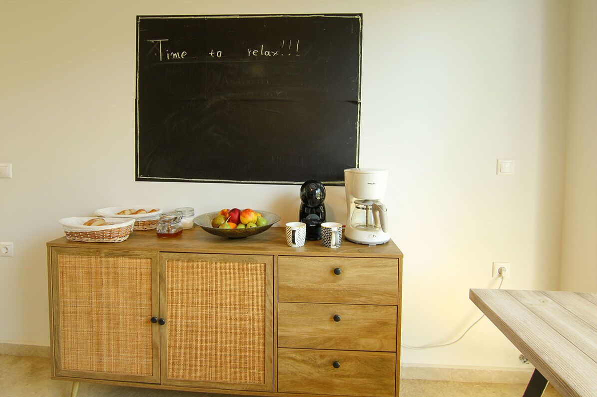 A kitchenette with a blackboard displaying "Time to relax!!!", a fruit bowl, coffee maker, and a basket of bread on a wooden cabinet.