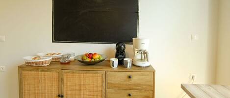A kitchenette with a blackboard displaying "Time to relax!!!", a fruit bowl, coffee maker, and a basket of bread on a wooden cabinet.