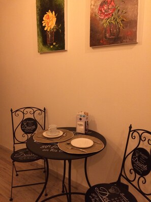 Una mesa redonda pequeña con dos sillas, cada una con un mantel individual con motivos de postres. Encima de la mesa, hay dos cuadros de flores.