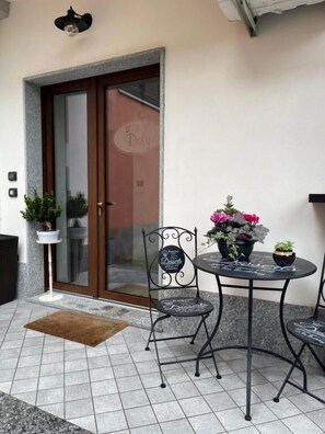 Un espacio al aire libre con una mesa y dos sillas, una puerta con panel de vidrio y plantas en macetas.